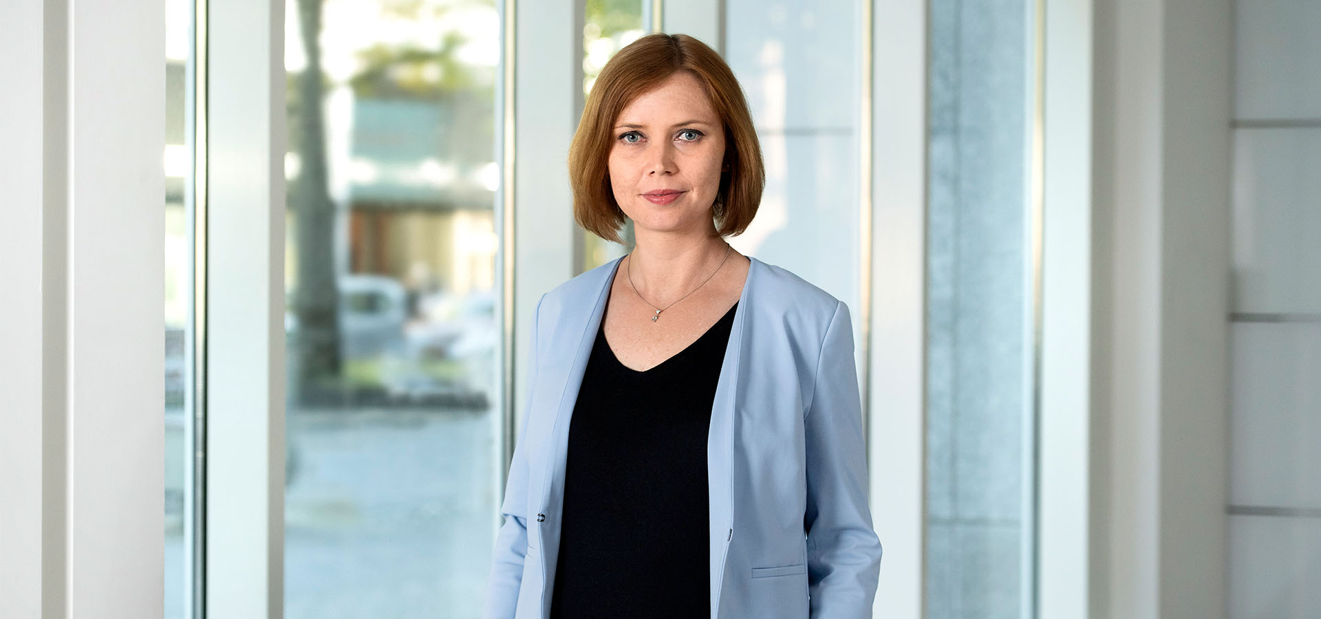 Eine Frau in schwarzer Bluse und blauer Jacke steht vor einem großen Fenster.