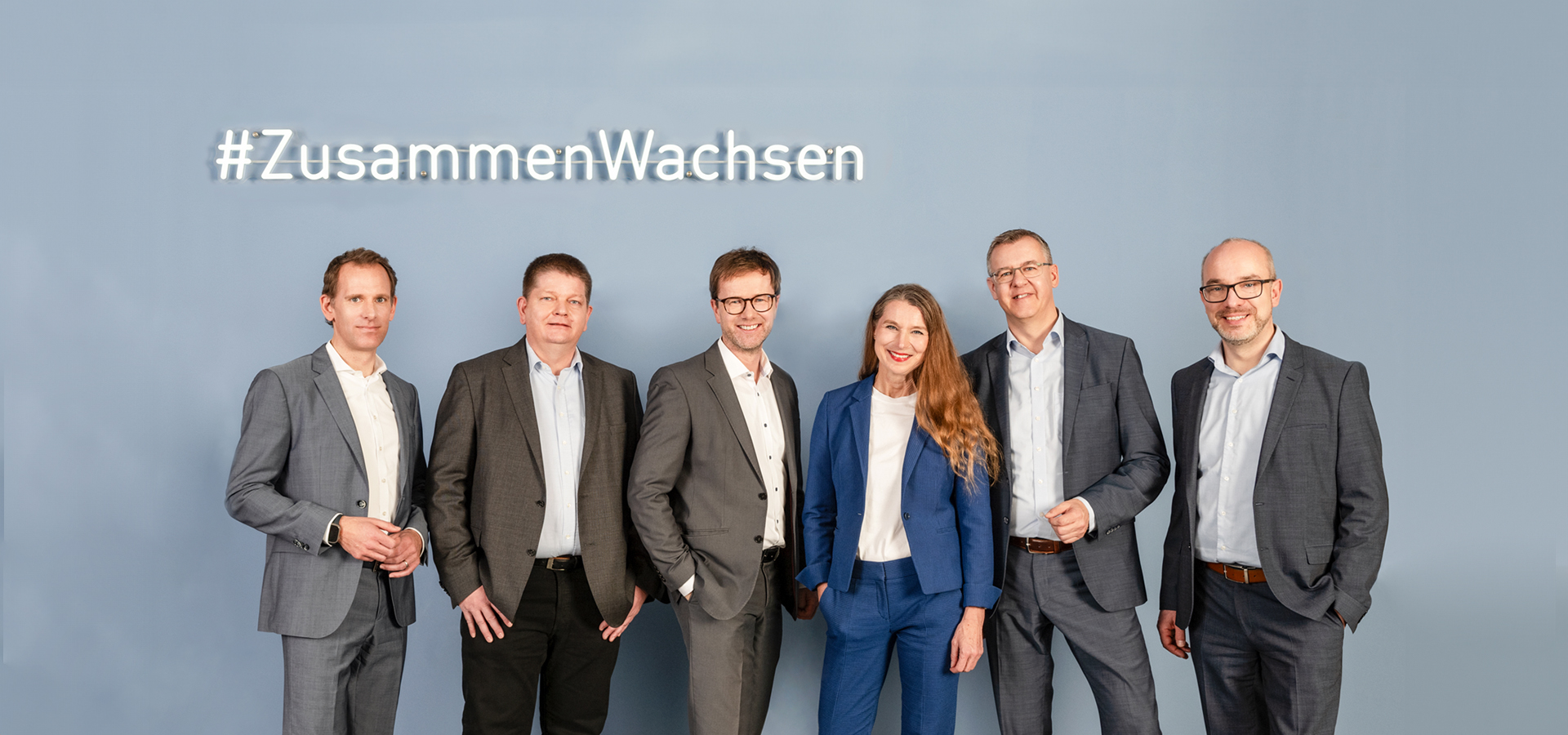 Fünf Männer und eine Frau stehen vor einer grauen Wand und dem Leuchtschild mit der Aufschrift #ZusammenWachsen