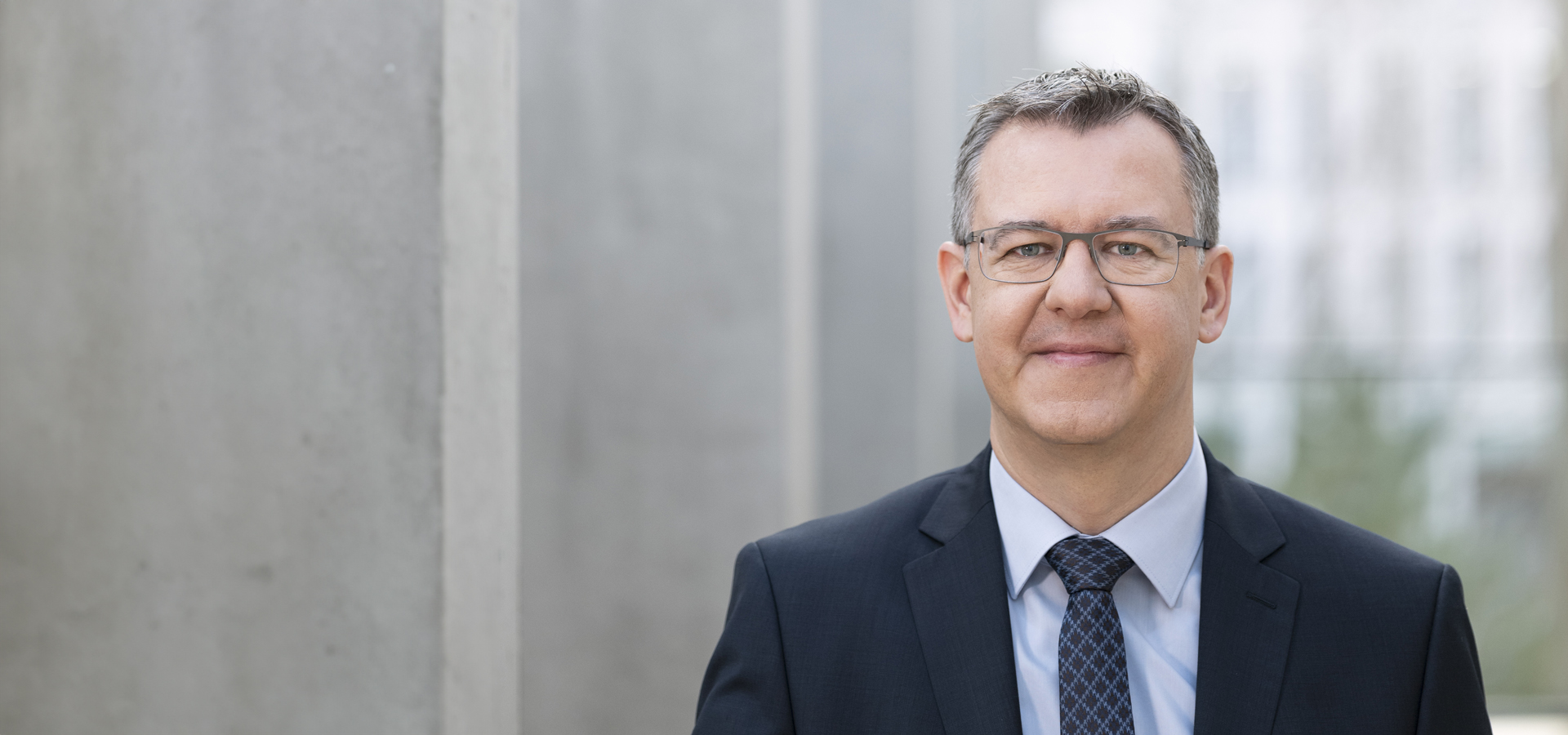 Ein Mann in Brille und Anzug steht vor einer grauen Betonwand.
