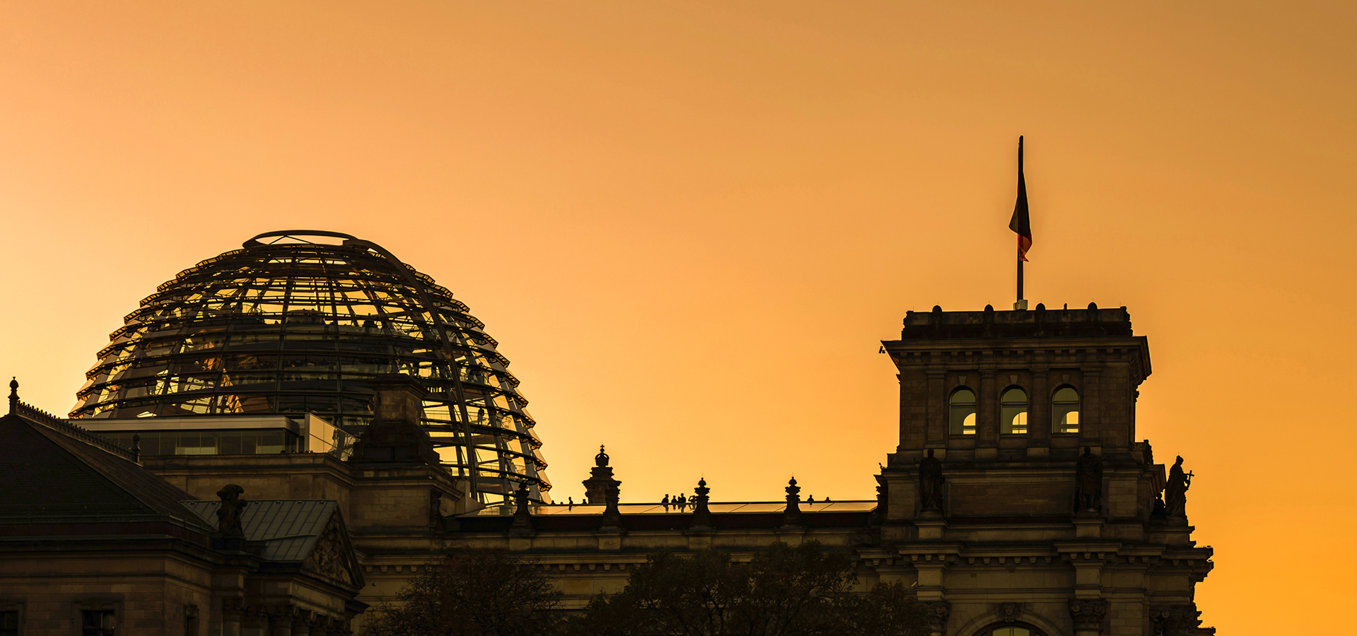 Reichstagsgebäude in Berlin bei Sonnenuntergang, mit der markanten Glaskuppel und deutscher Flagge auf dem Turm. Symbolbild für politische Prozesse und rechtliche Rahmenbedingungen wie den Normenvertrag.