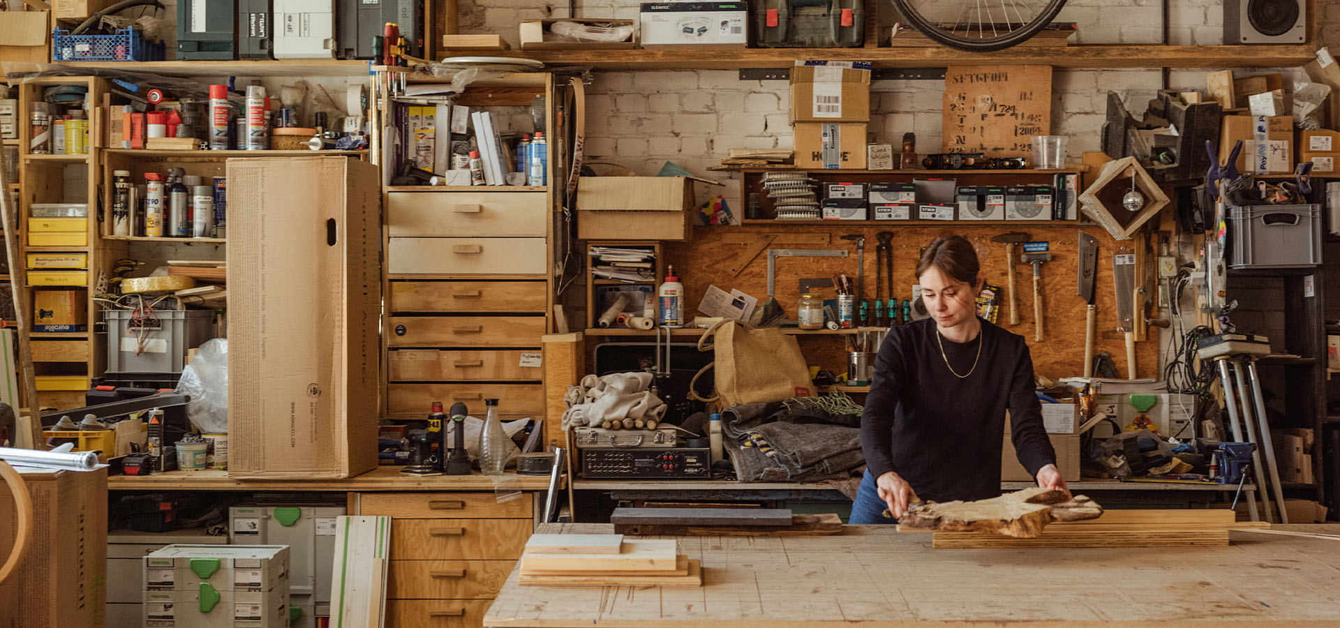 Geschäftsführerin Christina Reimann Holz-Instituts arbeitet in einer gut gefüllten Schreinerwerkstatt an einem großen Stück Holz auf einer Werkbank, umgeben von Werkzeugen, Maschinen und Holzzuschnitten